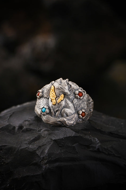 A sculpted silver-tone fox ring with gold-tone details and small blue-green and red accents, resting on dark stone.