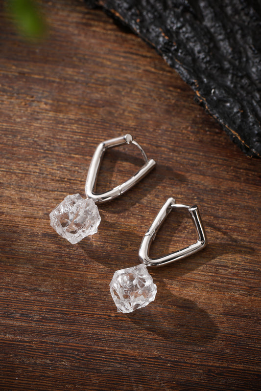 Two irregular clear quartz drops attached to angular silver-colored hoops, arranged diagonally on brown wood.